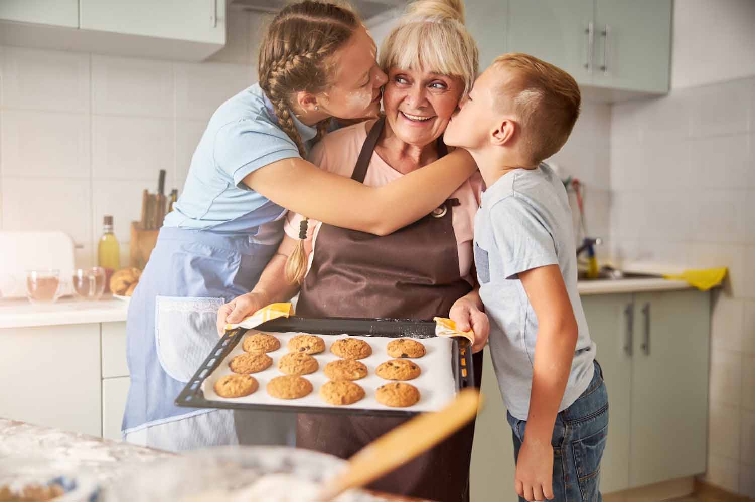 Blissful grandma holding a pan of fresh baked cookies, being kissed by thankful grandchildren