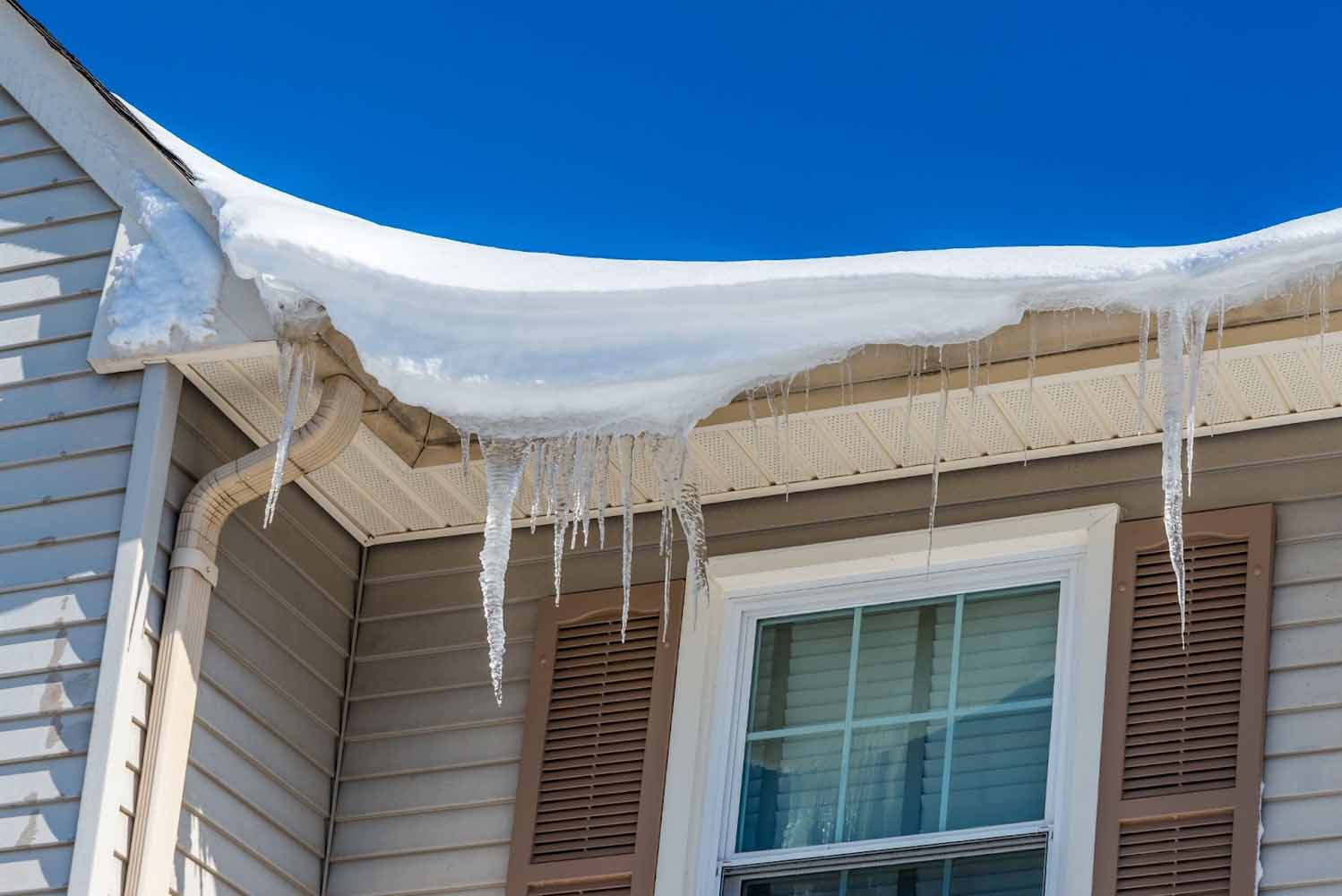 Ice dam, icicles, and snow built up on the eaves of a tan house