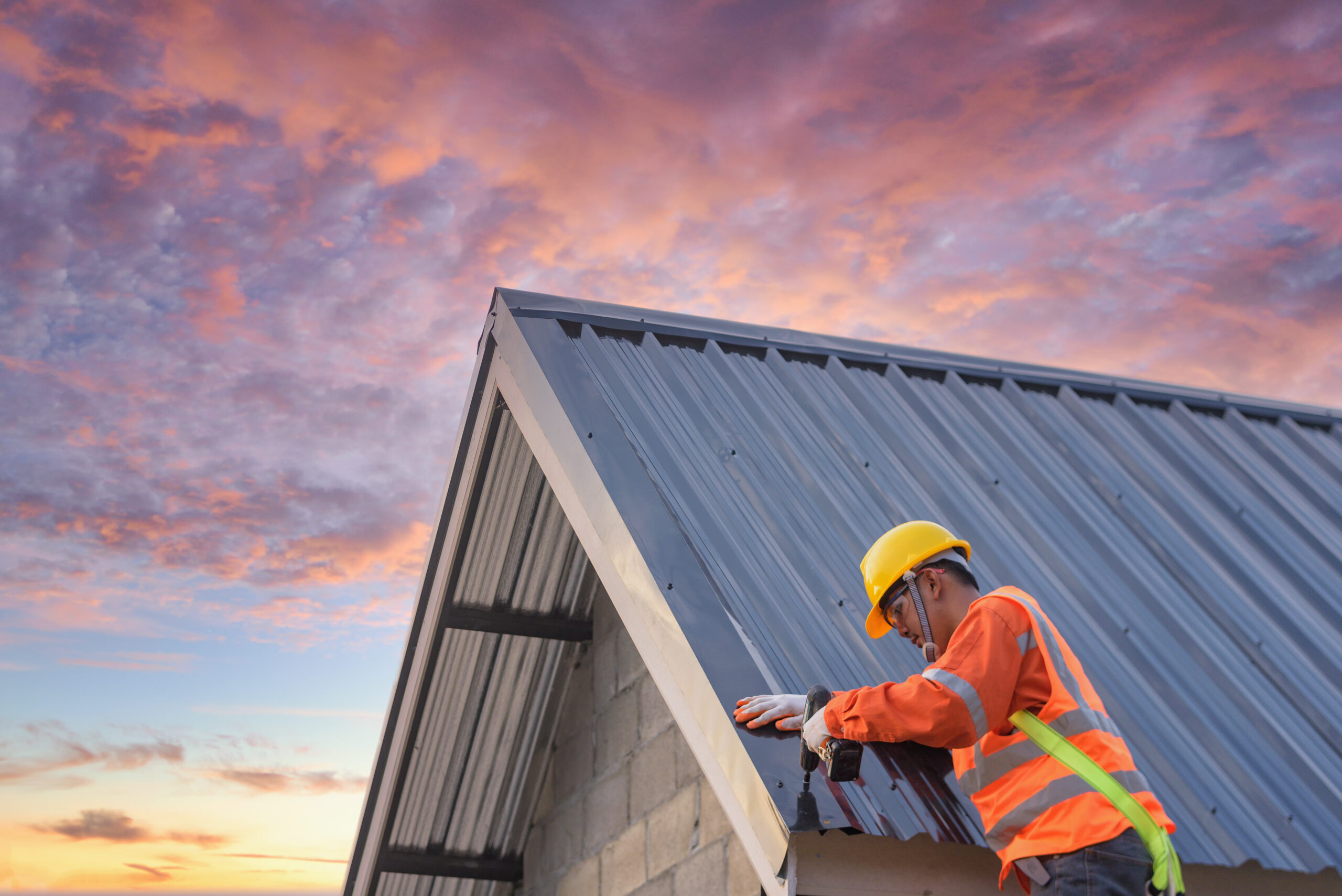 Roofer in hardhat and safety vest installs metal roof at sunset