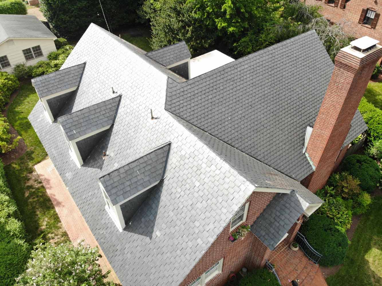 Aerial view of a large brick house with a complex gray shingle roof and multiple dormers.