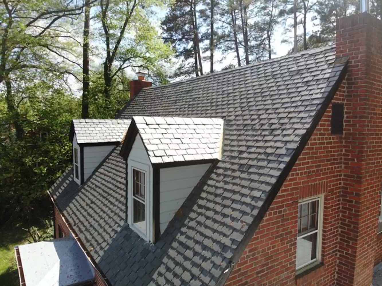 Aerial view of a brick house with multiple gray slate roofs and dormer windows surrounded by trees. 