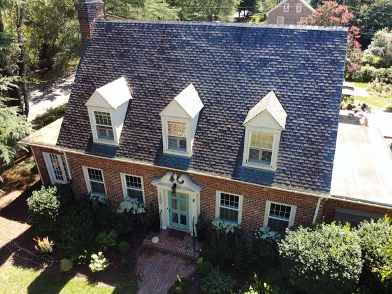 Aerial view of a classic brick house with a blue shingle roof and three dormer windows surrounded by greenery. 