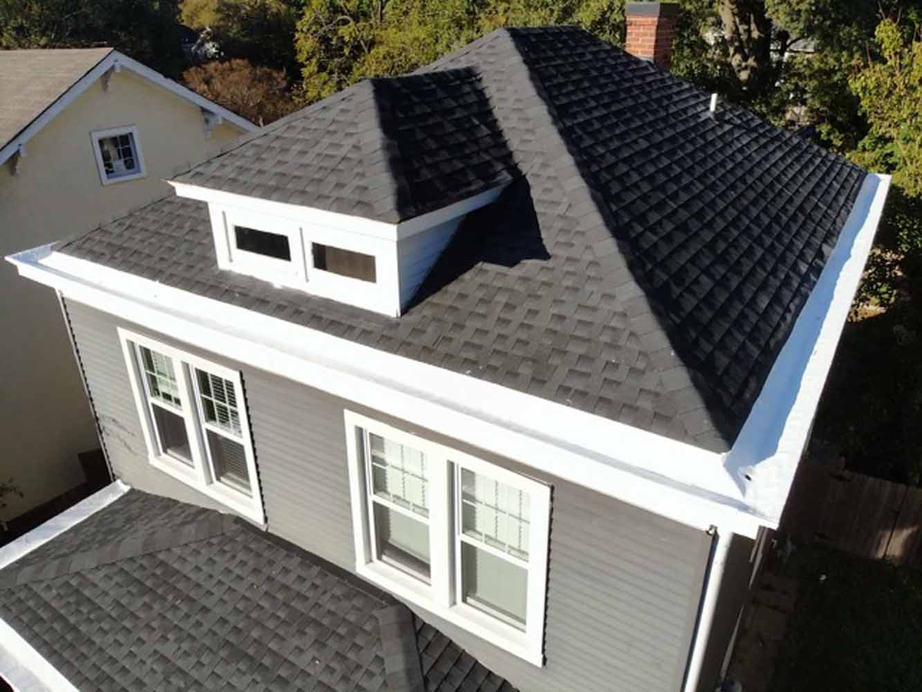 Aerial view of a modern house with dark gray shingles, featuring a prominent dormer window and white trim. 