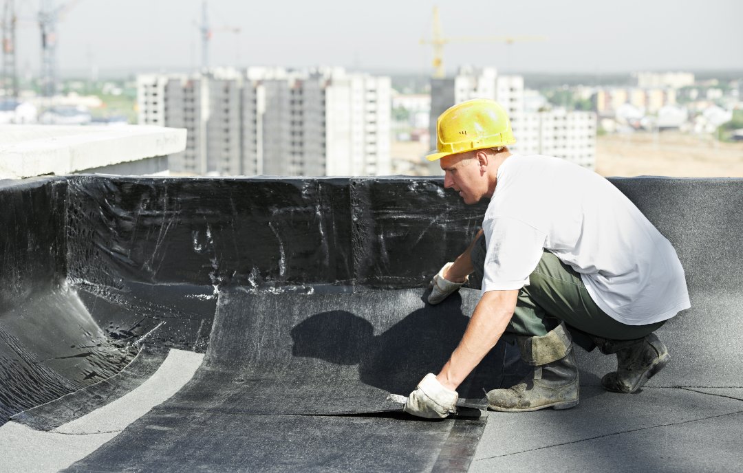 Worker in a hard hat and gloves applies waterproofing material to a rooftop with city skyline in background. 