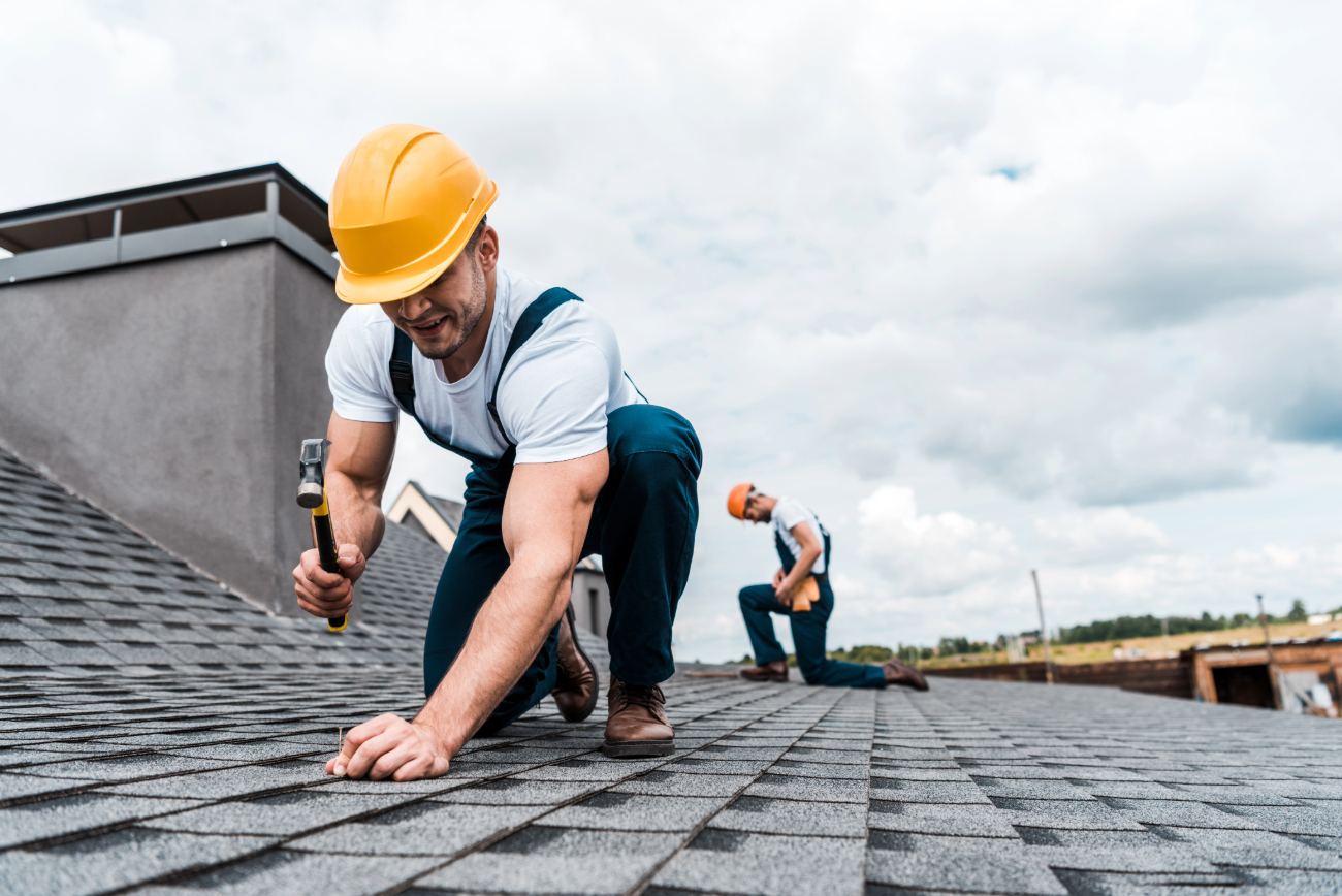 Two roofers in hard hats and overalls working on a shingle roof, one hammering a nail. 