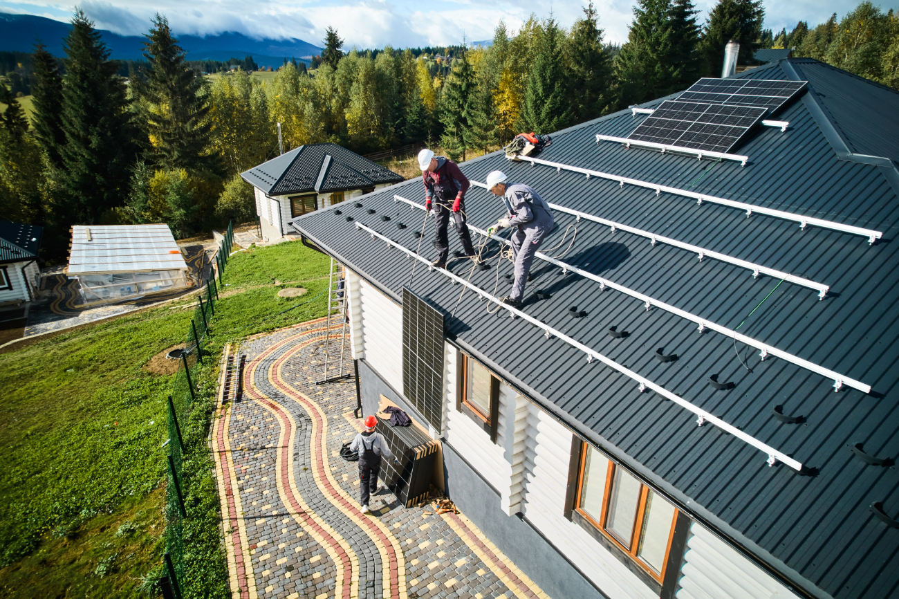 Workers installing solar panels on a house roof with mountain and forest backdrop. 