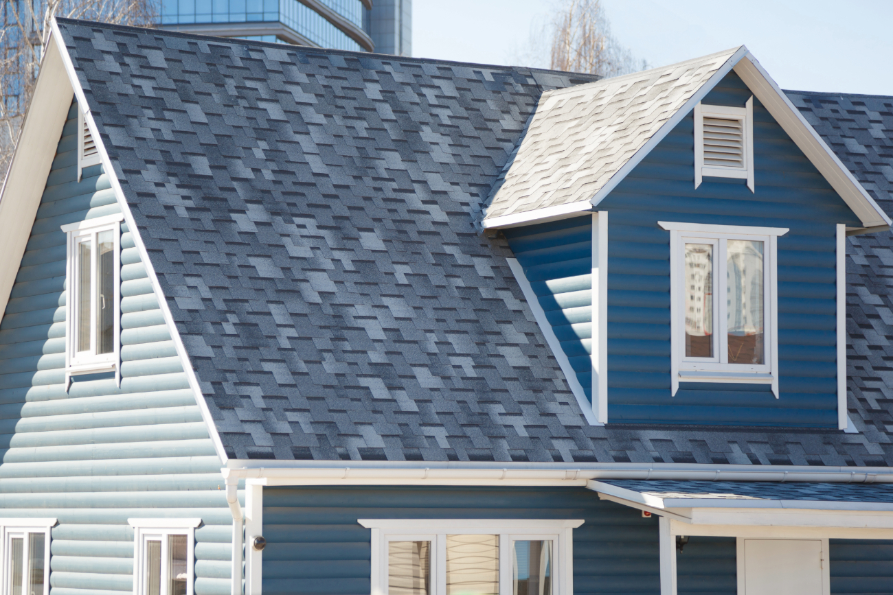 Close-up of a blue house's roof featuring contrasting dark gray shingles and a small attic window. 