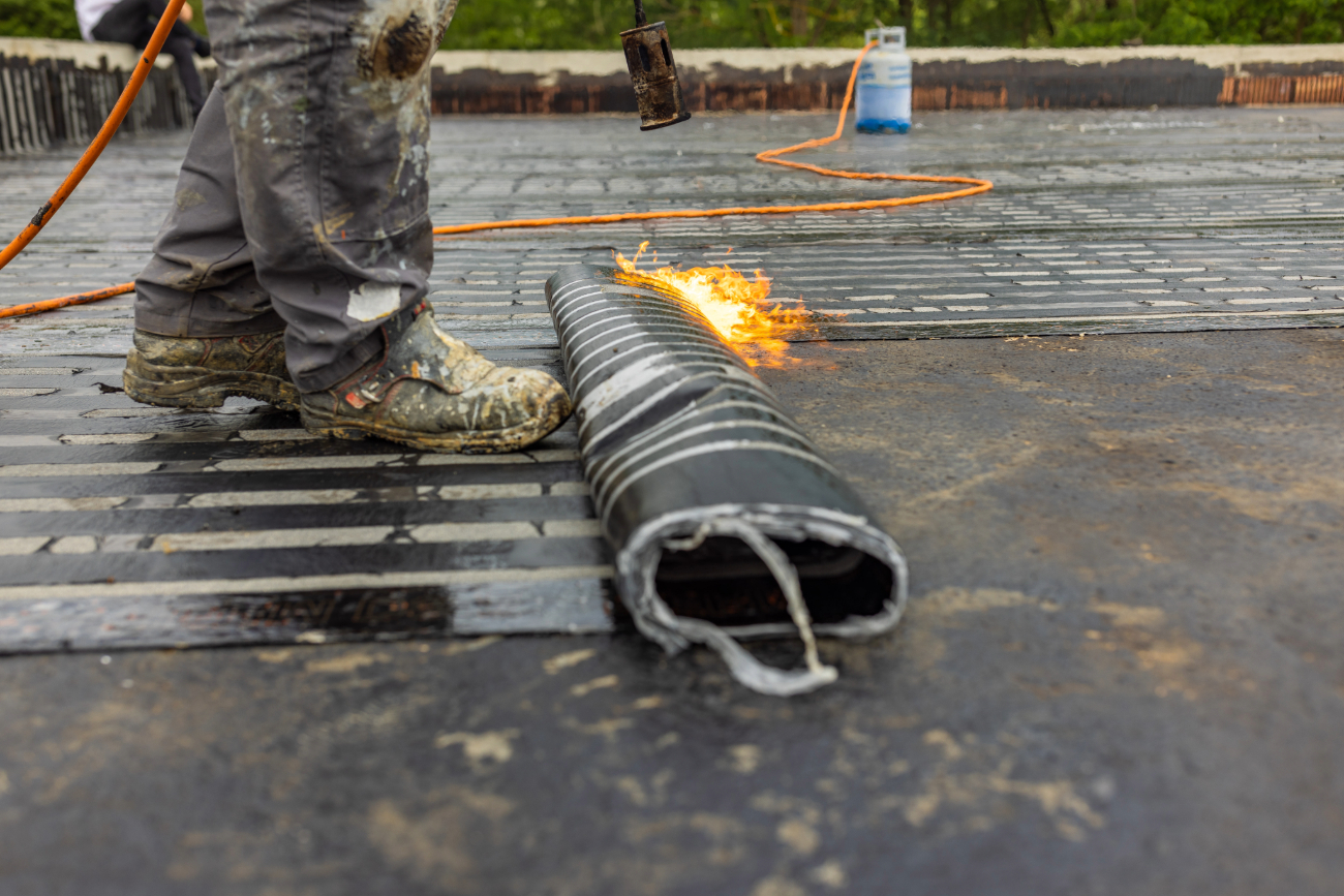 Roofer using a torch to seal a membrane on a flat roof, with tools and equipment visible. 