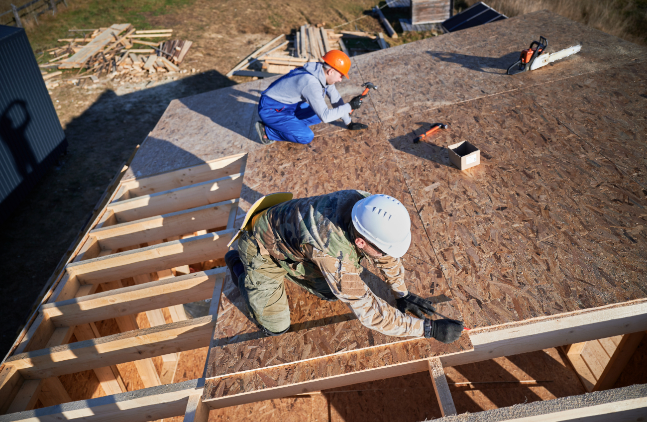 Construction workers in helmets installing plywood sheathing on a new roof frame at a construction site. 