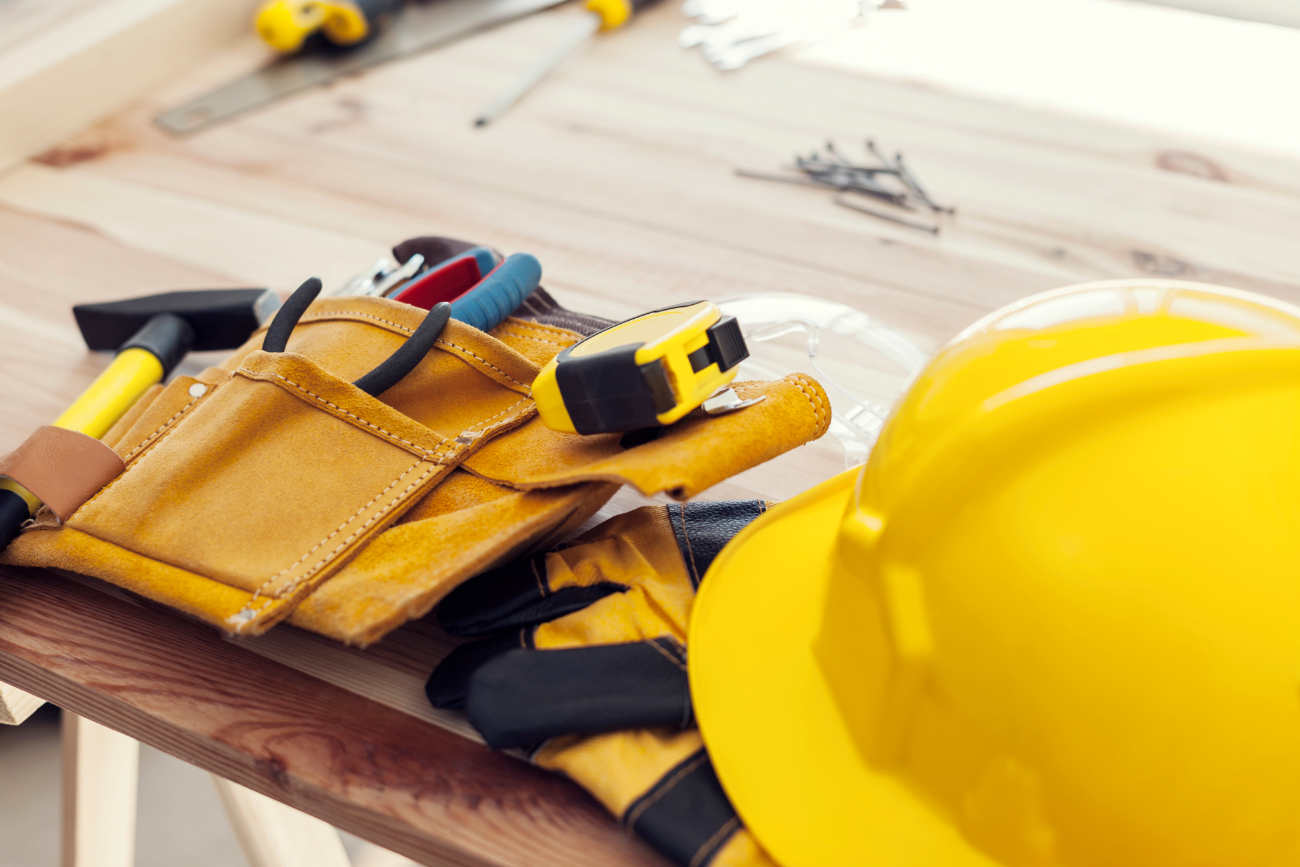Close-up of a construction toolkit with a yellow hard hat, measuring tape, and various tools on a wooden surface. 