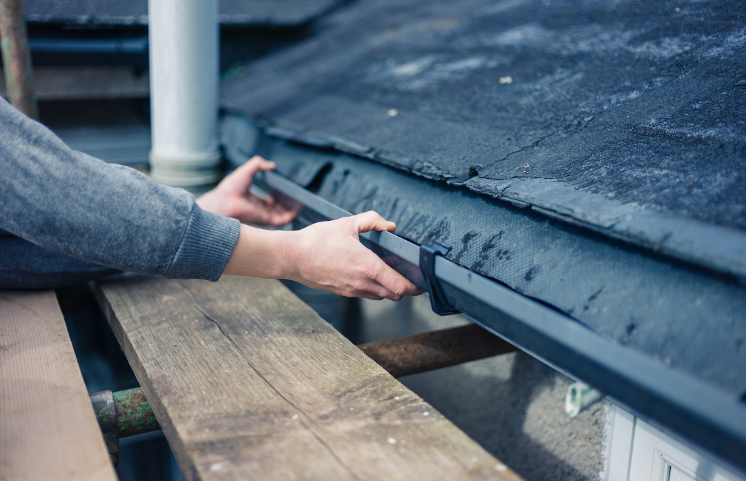 Person adjusting a gutter on a tar-covered roof edge near a white column and wooden bench. 