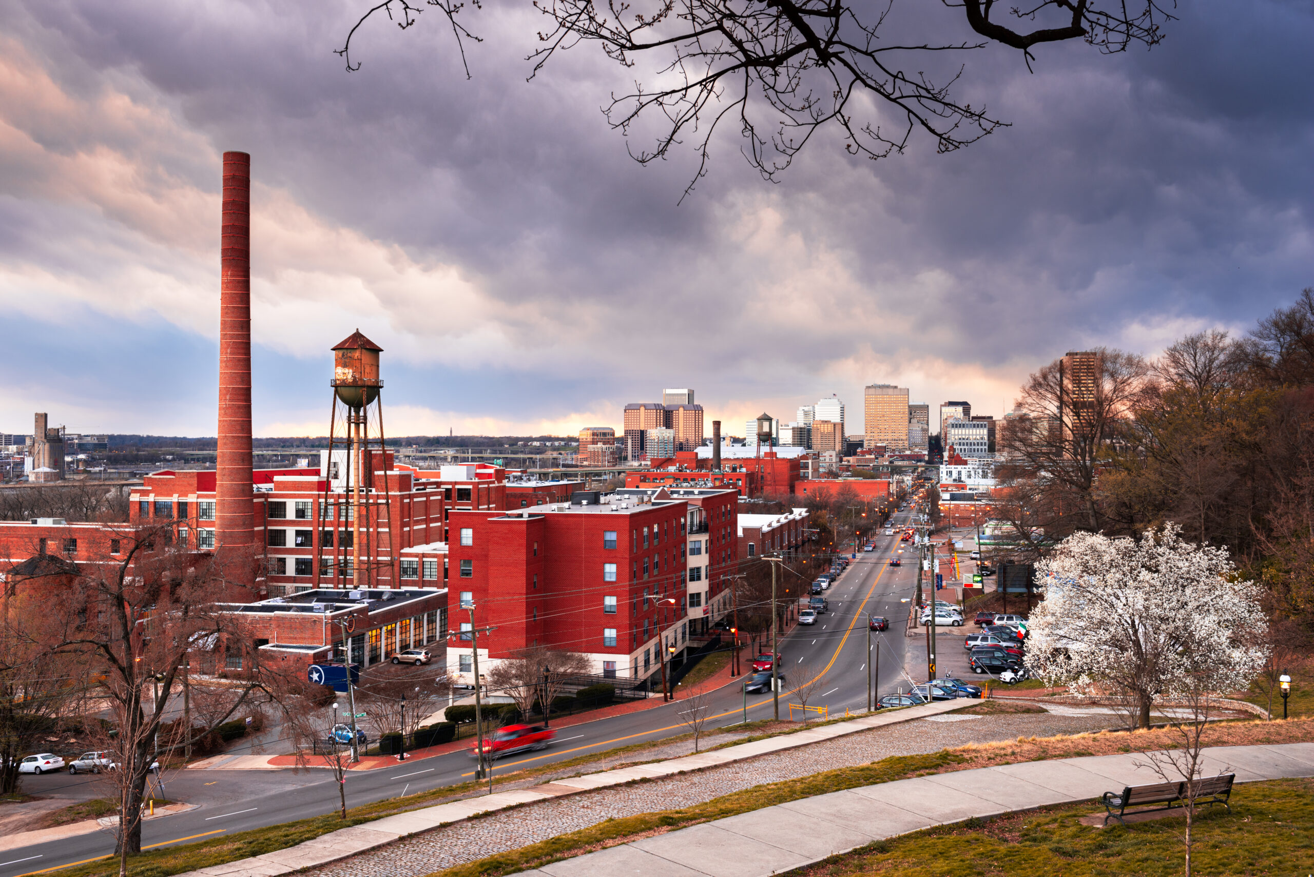 Sunset over a cityscape featuring a historic smokestack and water tower with modern buildings in the background. 