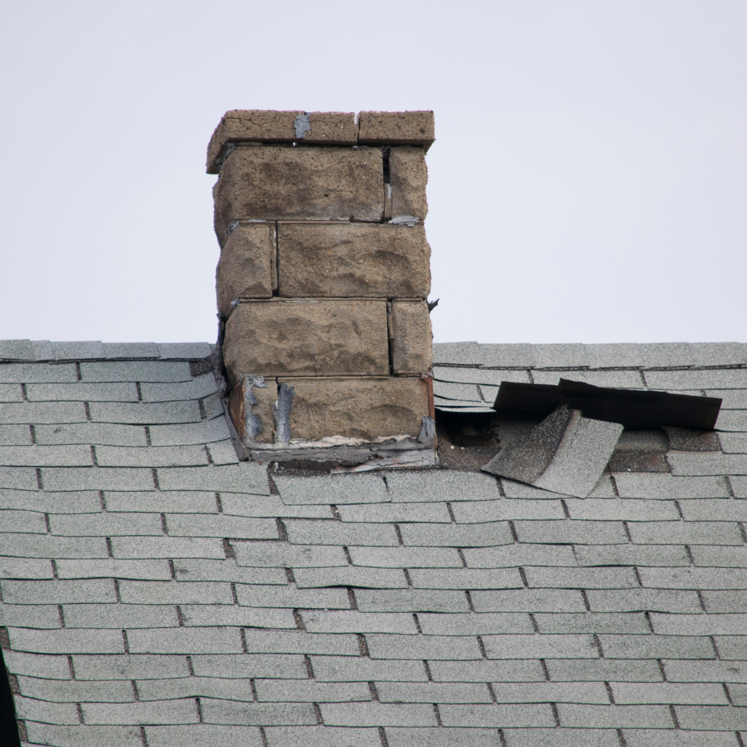 Close-up of a damaged chimney with missing bricks on a house with a gray shingle roof. 