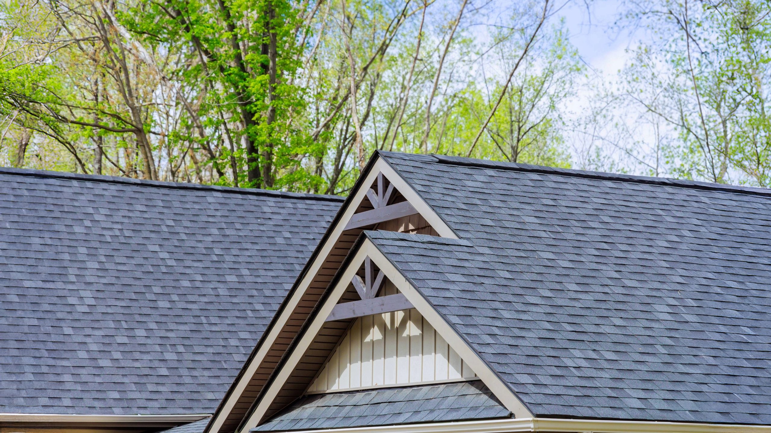 Close-up of a residential roof with dark gray asphalt shingles, featuring multiple roof slopes and decorative gable accents, surrounded by green spring trees in Richmond, VA.