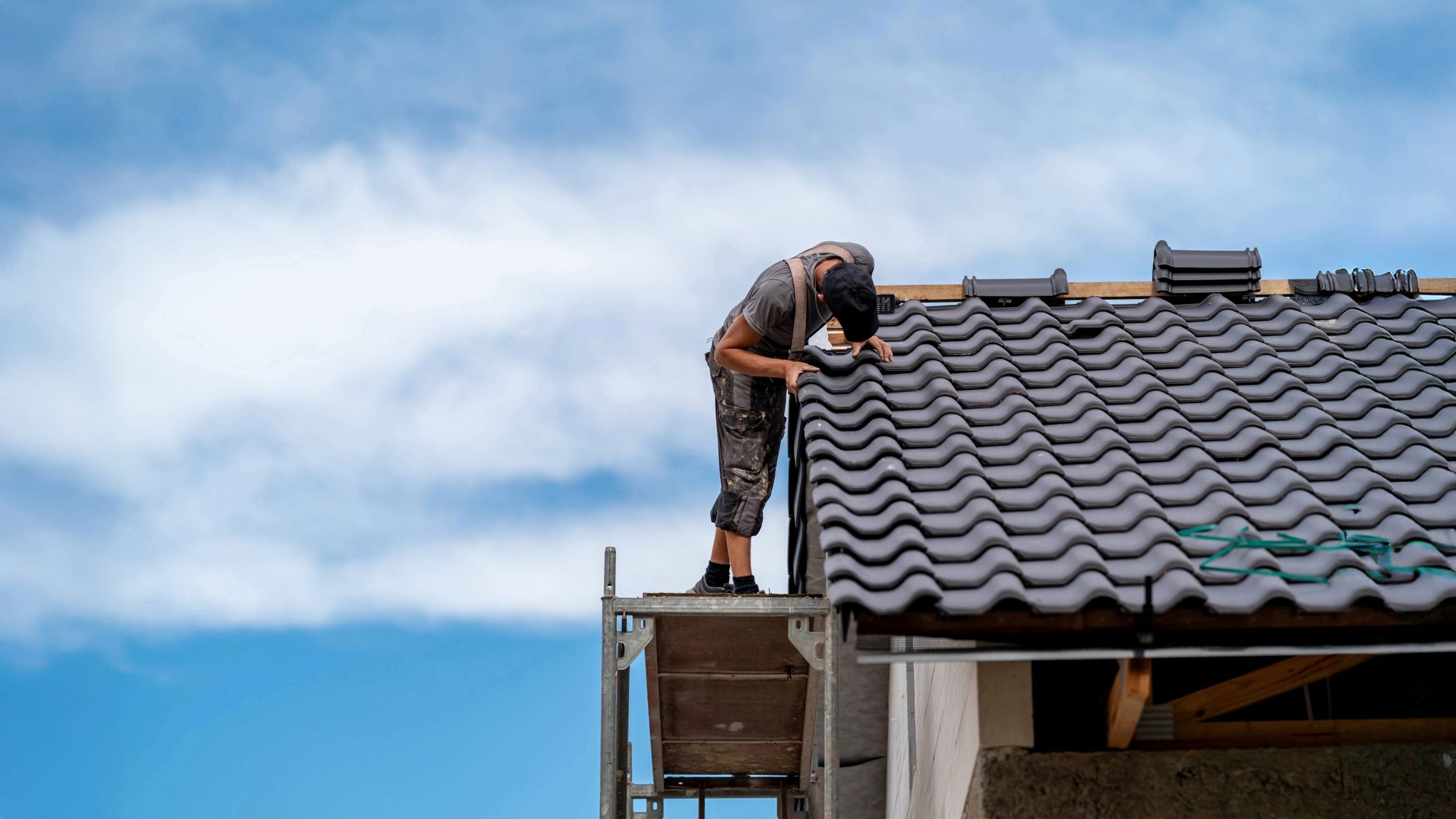 Roofer standing on scaffolding, inspecting and repairing gray tile roofing under a blue sky — representing professional roofing repair services.