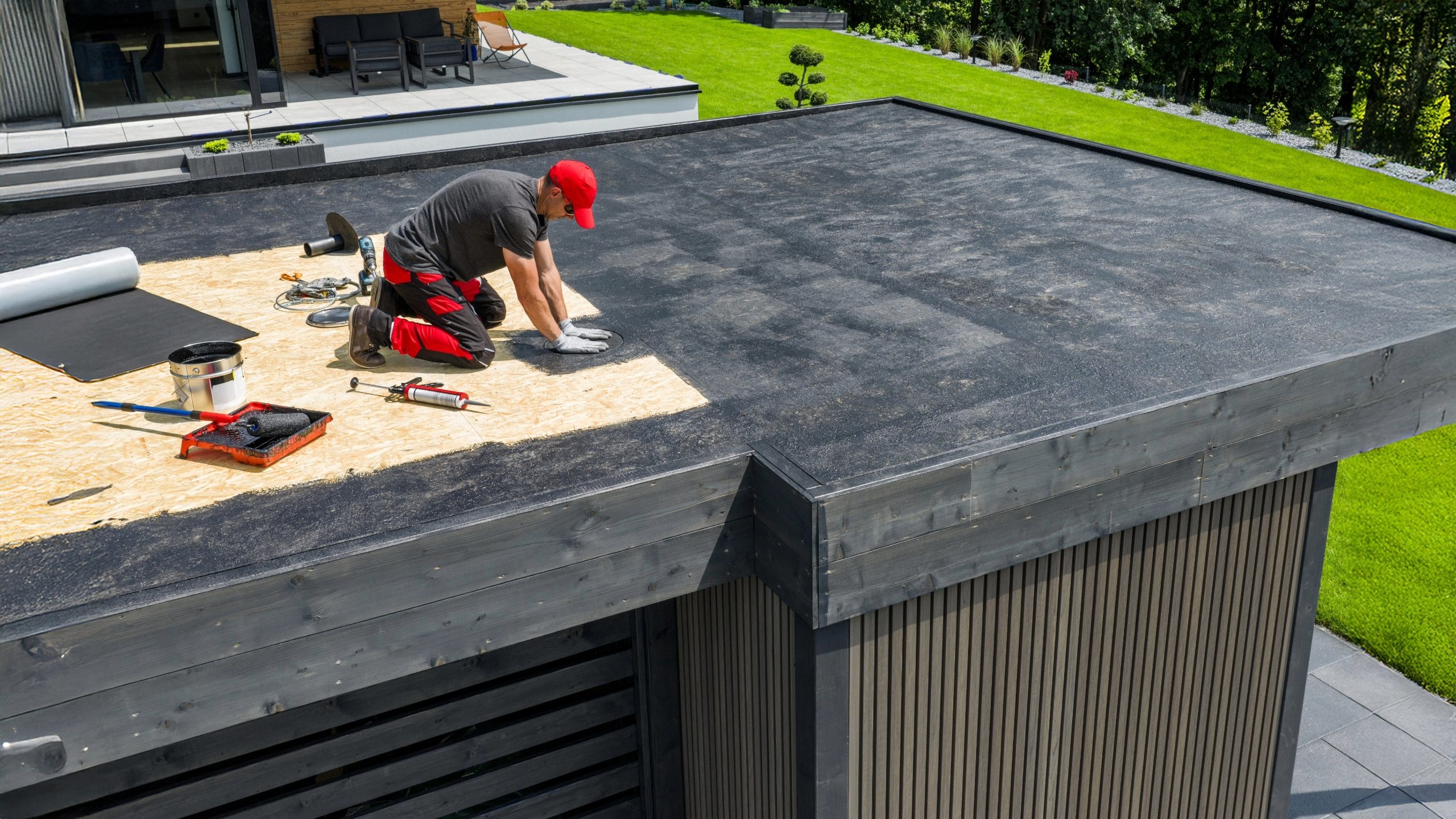 A roofing contractor installs a new flat roof membrane on a modern building using tools, adhesive, and roofing cement, with partially covered plywood and finished black roofing material visible.