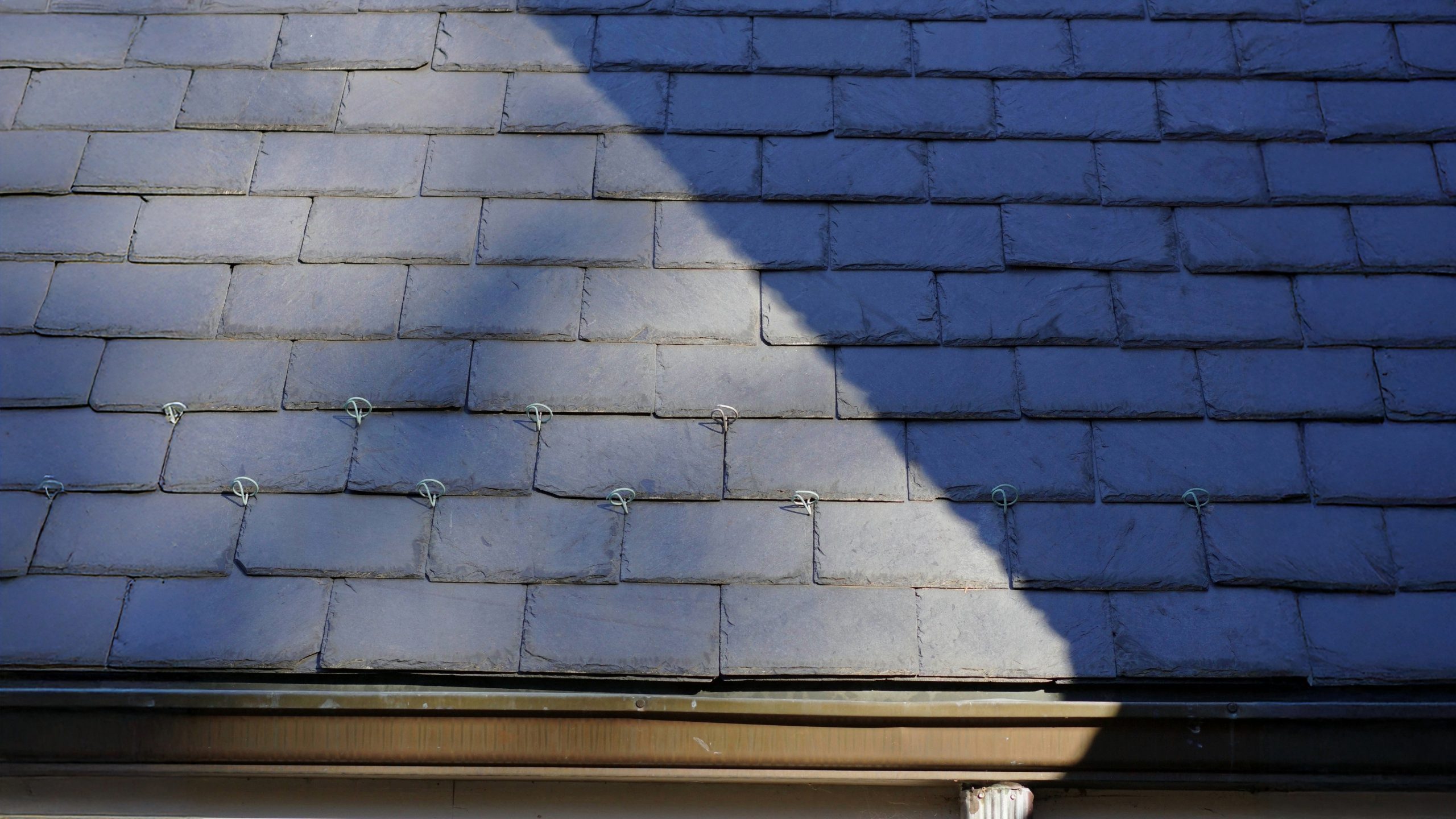 Close-up view of a slate roof with metal hooks securing individual slate tiles, partially shaded by a diagonal shadow, above a copper or metal gutter system.